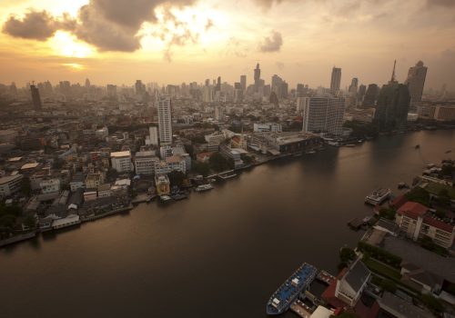 A skyline of central Bangkok taken from a high angle. There is a yellow sun and the city is still cast in some shadow.