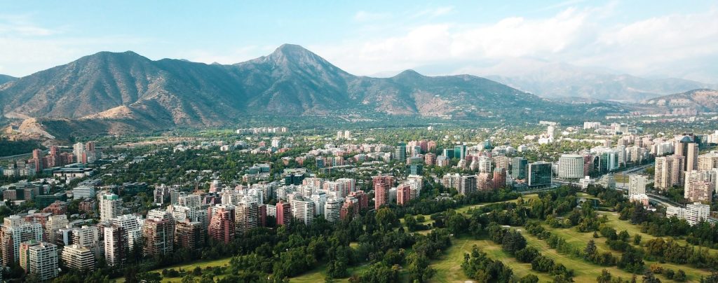 Image of city beneath mountains with green space in foreground. The sun is bright.