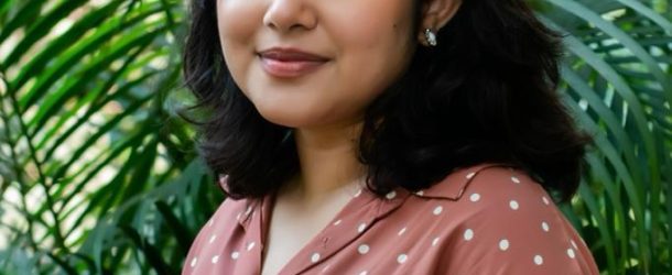 Image of Chief Heat Officer in Dhaka, Bushra Afreen. She wears a brown blouse with polka dots and stands in front of a green, leafy backdrop.