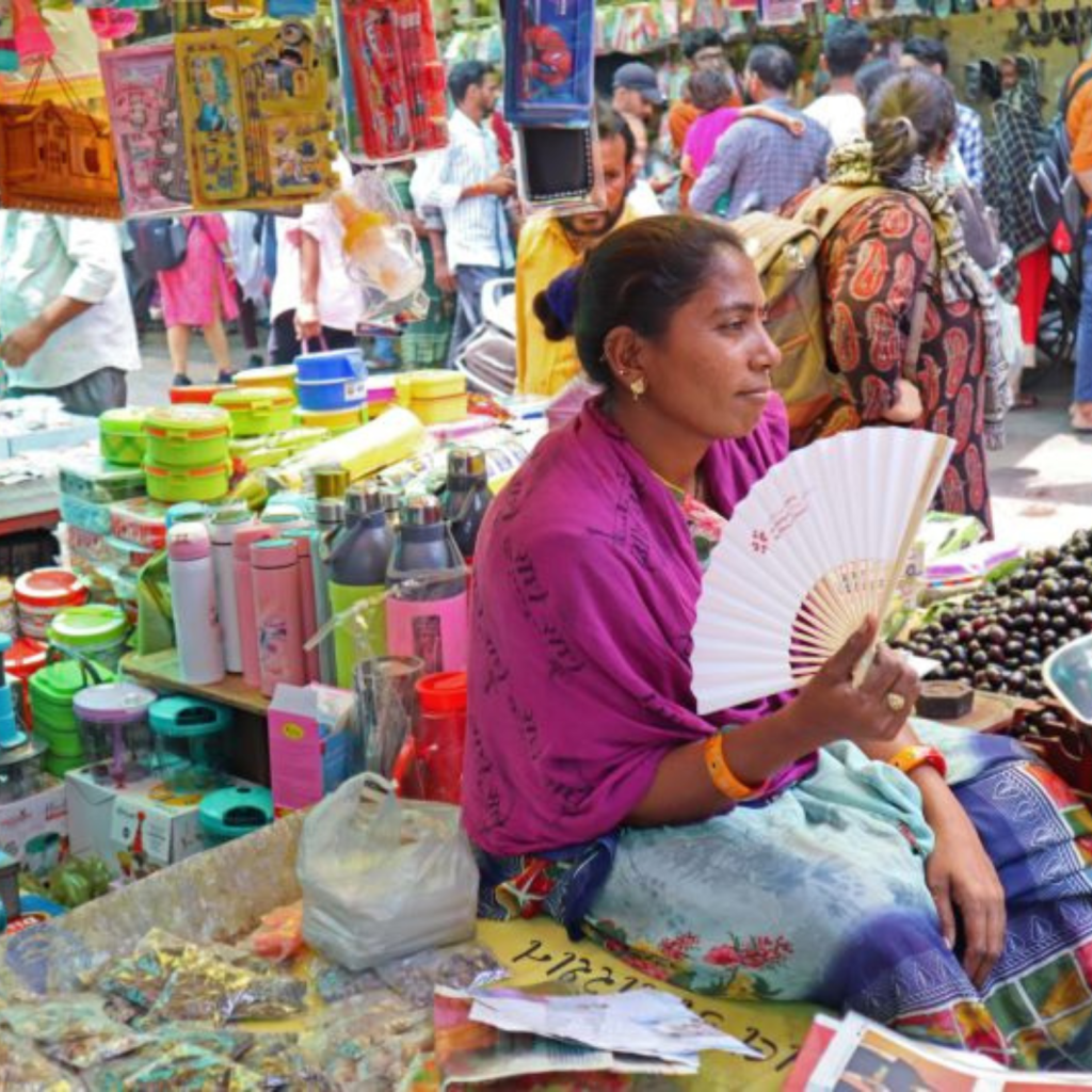 Image of a woman using a fan to cool herself in an outdoor market.