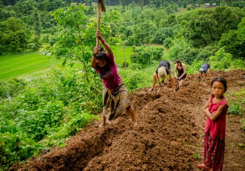 Image of local farmer tilling earth while children look on.